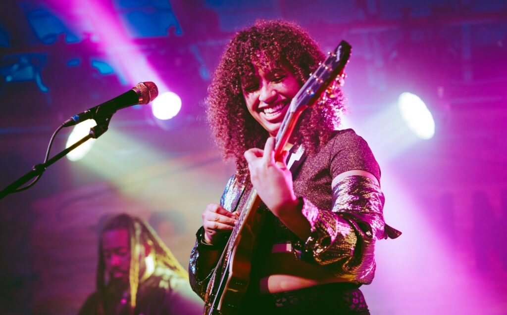 Jackie Venson smiles while playing guitar on stage under colorful lights during a live performance.