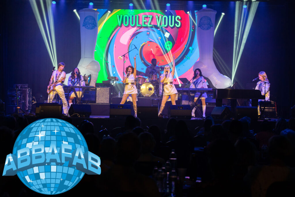 ABBAFAB performs live at New Spire Arts in Frederick, MD, with colorful lights and the word “Voulez Vous” displayed on screen during their high-energy ABBA tribute concert.