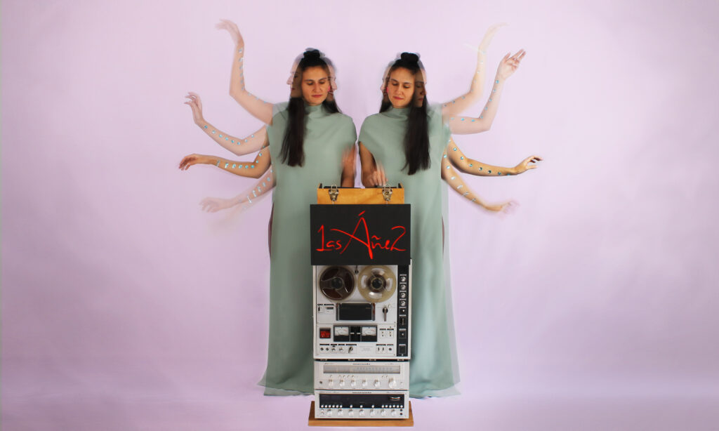 Las Áñez twin musicians performing beside a reel-to-reel tape machine against a soft pink background.