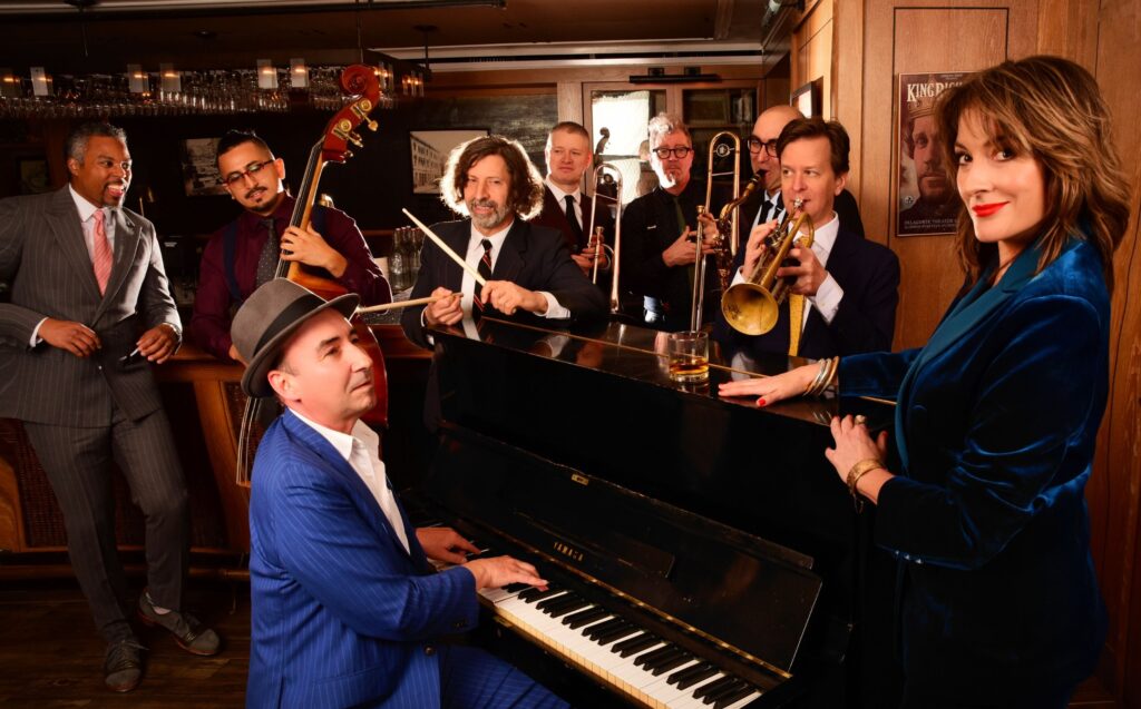 The Hot Sardines band gathered around a piano with horns, bass, and vocals in a vintage lounge setting.